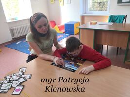 Pani Patrycja Klonowska siedzi z uczniem przy stoliku w klasie. Czytają sylaby. Na dole zdjęcia napis: mgr Patrycja Klonowska