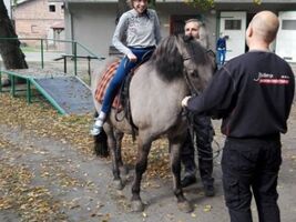 dziewczynka siedzi na grzbiecie konia o imieniu Hejnał, obok asekuruje hipoterapeuta, z przodu mężczyzna trzyma konia za wodze