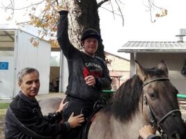 chłopiec siedzi na grzbiecie konia o imieniu Hejnał, unosi rękę w górę, obok asekuruje hipoterapeuta