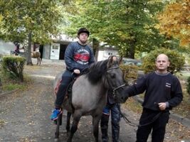 chłopiec siedzi na grzbiecie konia o imieniu Hejnał, obok asekuruje hipoterapeuta, z przodu mężczyzna trzyma konia za wodze